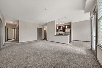 A spacious, empty room with a carpeted floor and a kitchen area in the background.